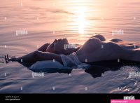 Silhouette of a young sexy nude woman with big beautiful breasts lies in  the waves of the reservoir in the rays of the setting sun Stock Photo